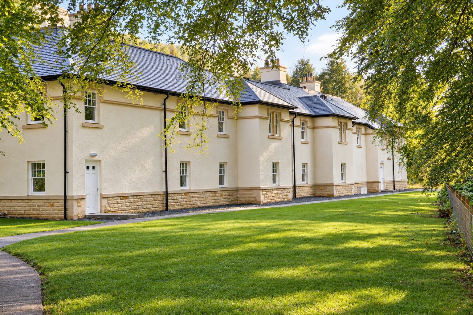 Long two-storey building finished in cream render over external wall insulation, with a stone base, seen across a lawn