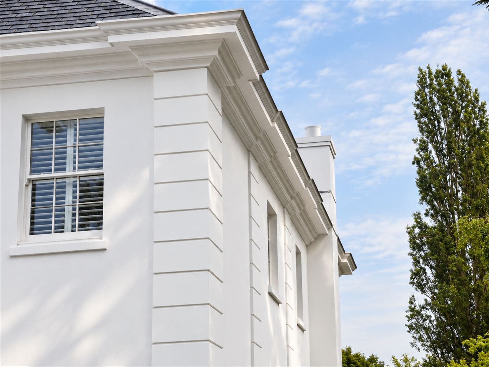 Corner of a white rendered house with a projecting cornice, quoins and a sash window
